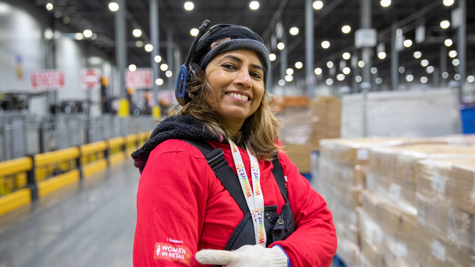 A female associate in a fulfillment center pointing to the sleeve on her red jacket, which says "Women in Retail"