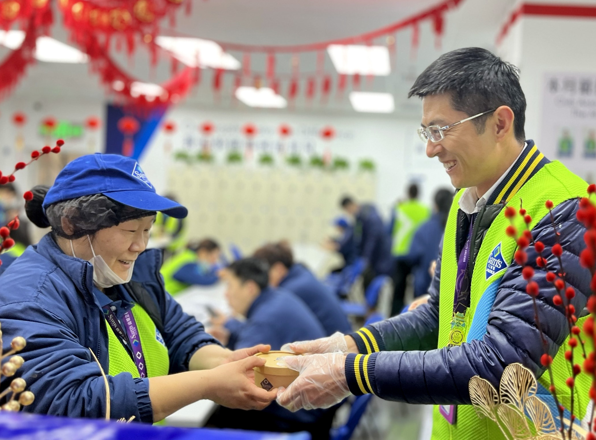 Two Sam’s Club associates smile as one gives a small container of food to the other. Red banners hang from the ceiling above.