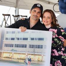 Brad Paisley and Kimberly Williams Paisley are holding a large poster featuring an architectural rendering of The Store in Nashville, TN. The setting appears to be an outdoor event under a tent, with chairs and decorations visible in the background. The poster prominently displays a modern building design with glass windows and a storefront.