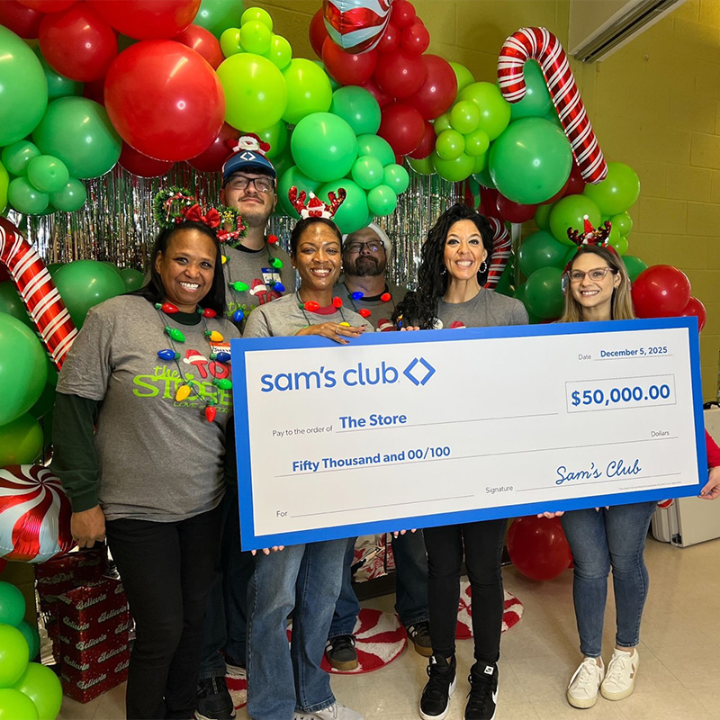 A group of Sam's Club volunteers stands in front of a festive holiday-themed backdrop featuring green and red balloons, candy cane decorations, and tinsel. They are holding a large donation check for $50,000. The check displays the name 'Sam's Club' prominently.