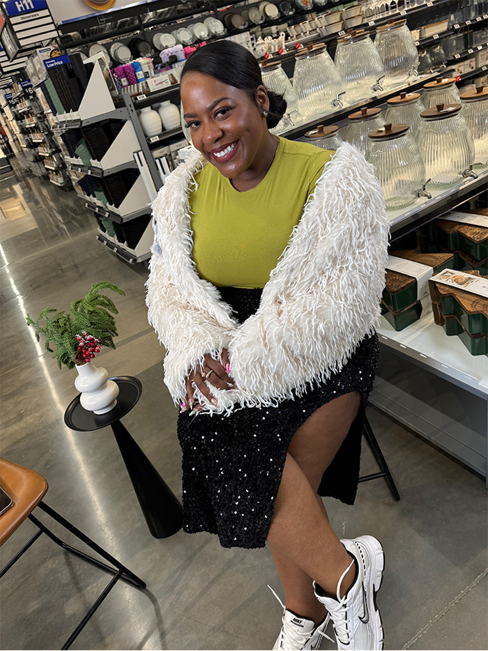 Walmart & Sam's Club Radio's Kirby Gwen, wearing a white textured jacket, lime green top, black sequined skirt and white sneakers, is seated in the Melissa, TX, Walmart Super Center. She is positioned near a small black table with a potted plant. The background features shelves with glass jars and home decor items.