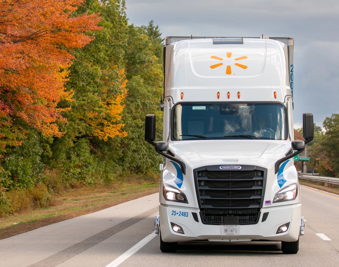A white Walmart semi truck is traveling down a highway bordered by trees with vibrant autumn foliage.