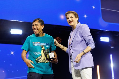 A Smiling Walmart Integrity in Action Award Recipient in a turquoise T-shirt stands on a brightly lit stage holding a small box-shaped award while smiling.