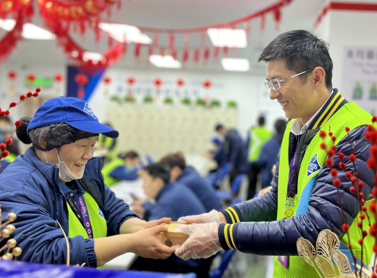 Two Sam’s Club associates smile as one gives a small container of food to the other. Red banners hang from the ceiling above.