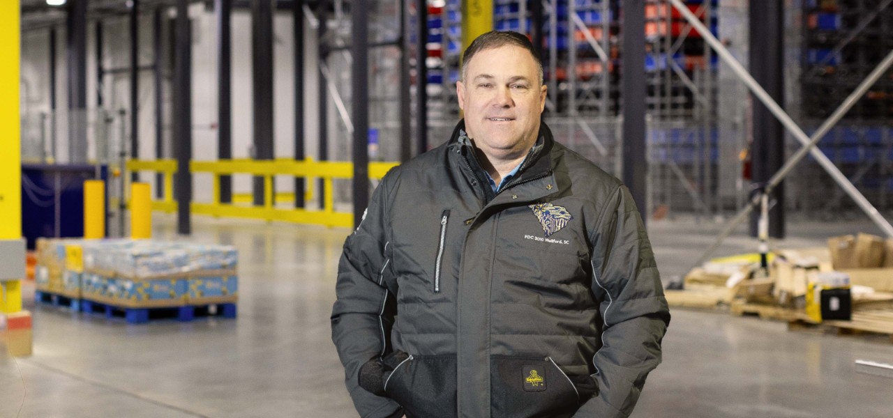 James Bright, a Walmart associate, wearing a blue vest and standing in a warehouse, is smiling at the camera.