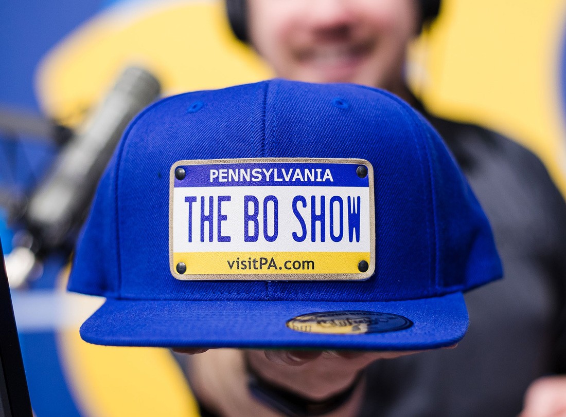 DJ Bo holds up a blue baseball cap with a decal of a Pennsylvania license plate that reads, "THE BO SHOW."